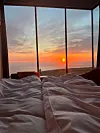 Utsikt til en rød solnedgang over hav og landskap sett fra ei seng gjennom et panoramavindu