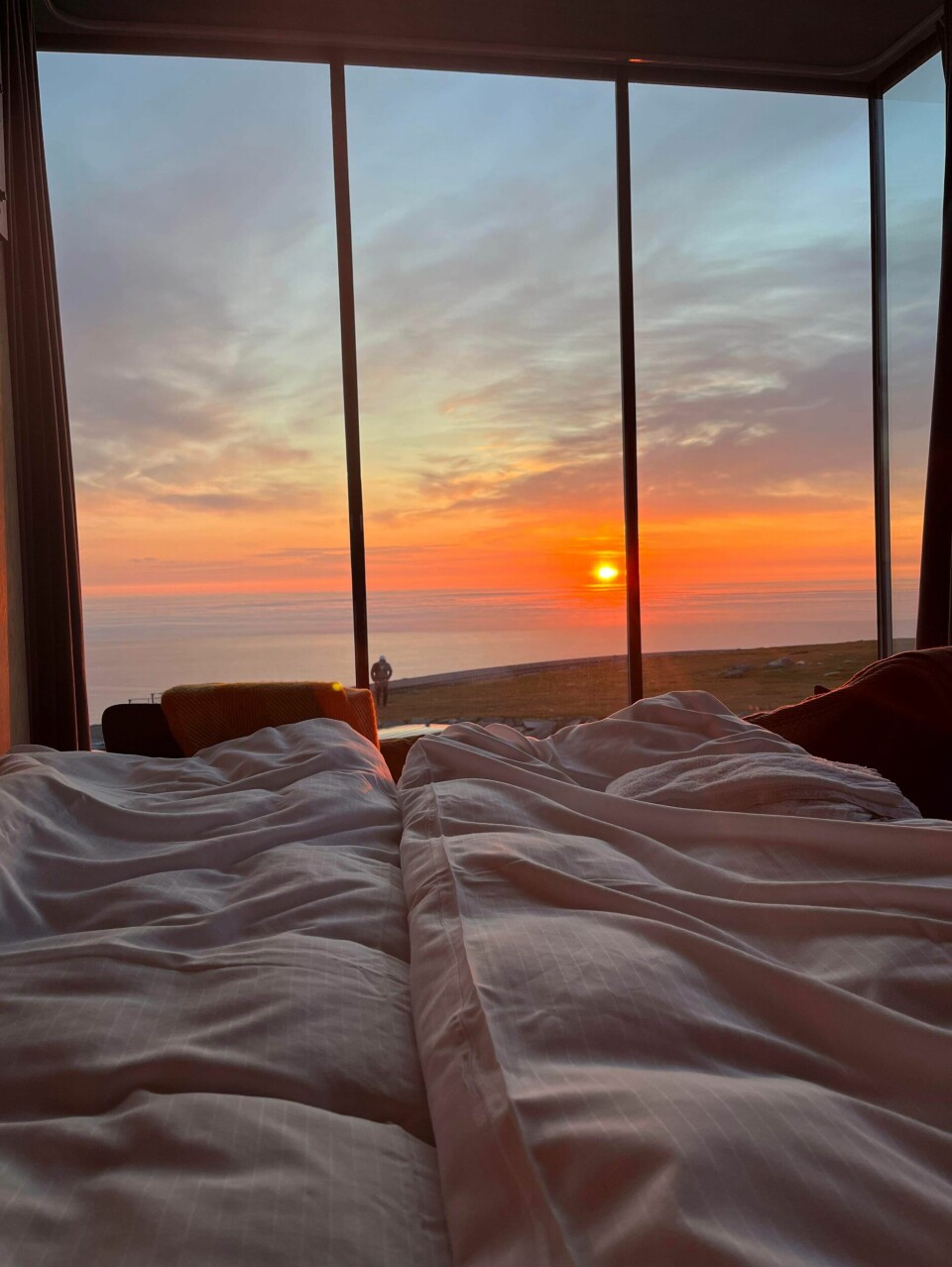 Utsikt til en rød solnedgang over hav og landskap sett fra ei seng gjennom et panoramavindu